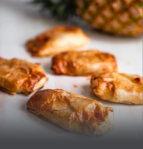 Pasteles hawaianos de hojaldre crujiente recién horneados, rellenos de jamón, queso y piña, acompañados de una piña natural al fondo en Cali.